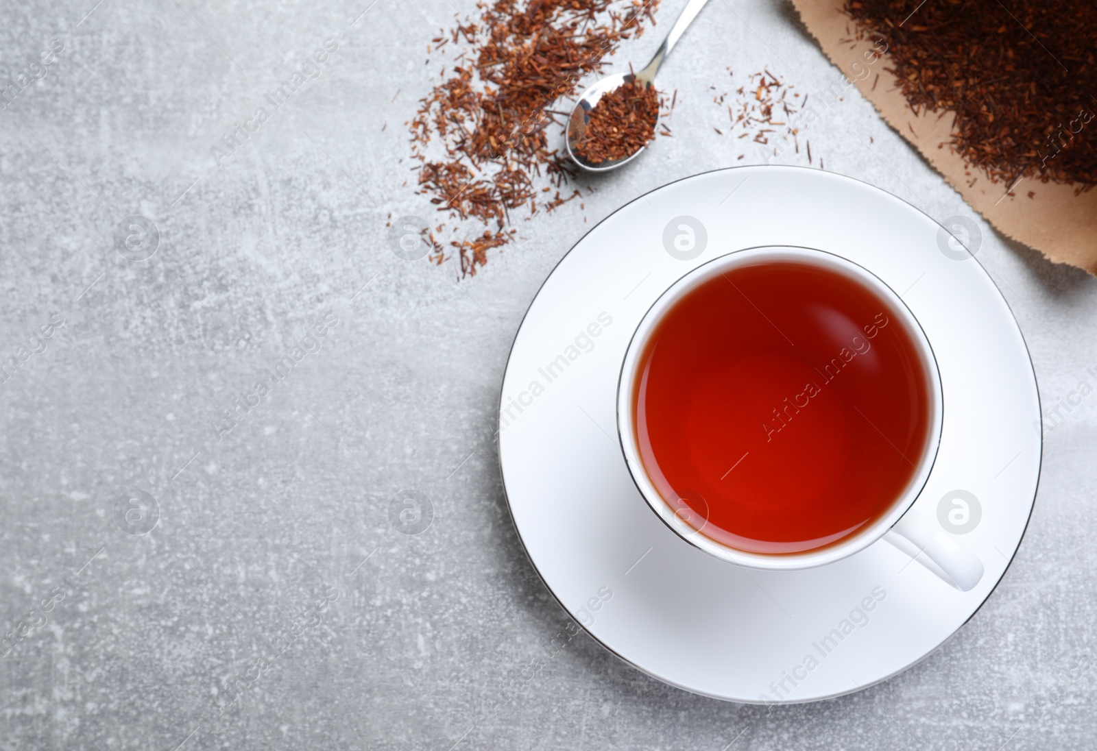 Photo of Ceramic cup of aromatic rooibos tea, spoon and scattered dry leaves on grey table, flat lay. Space for text