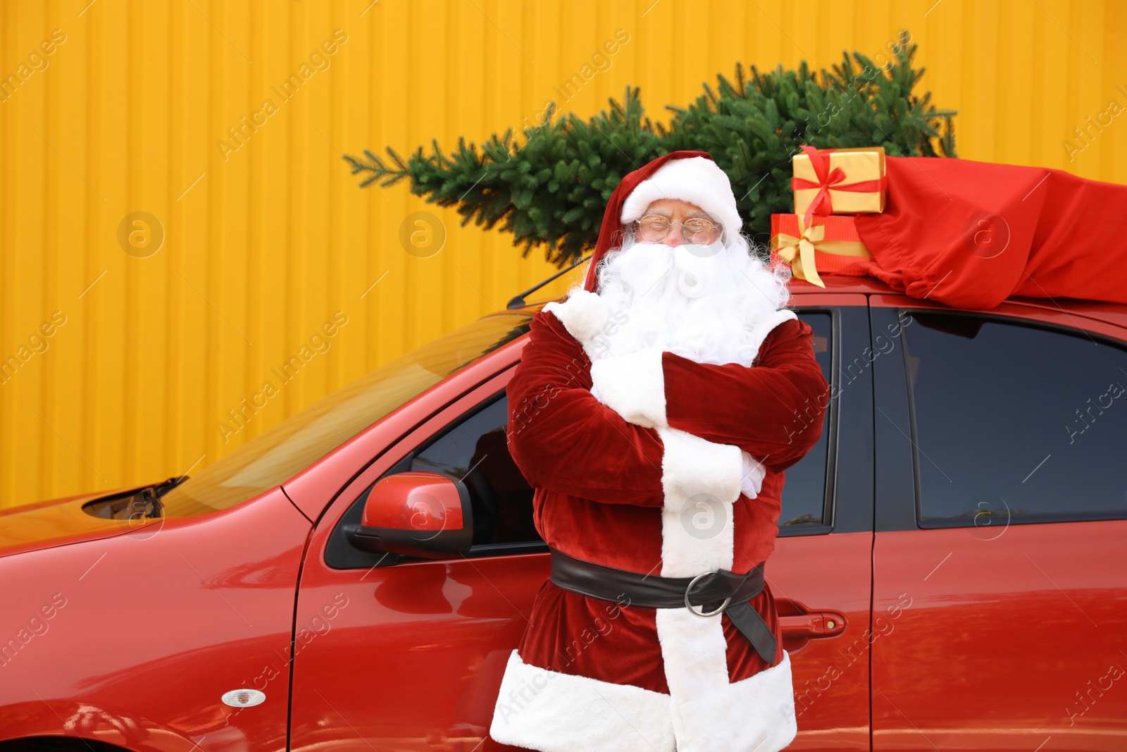 Photo of Authentic Santa Claus near car with fir tree and bag full of presents on roof against yellow background