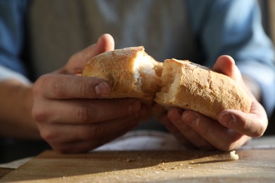 Photo of Man breaking loaf of fresh bread at wooden table indoors, closeup