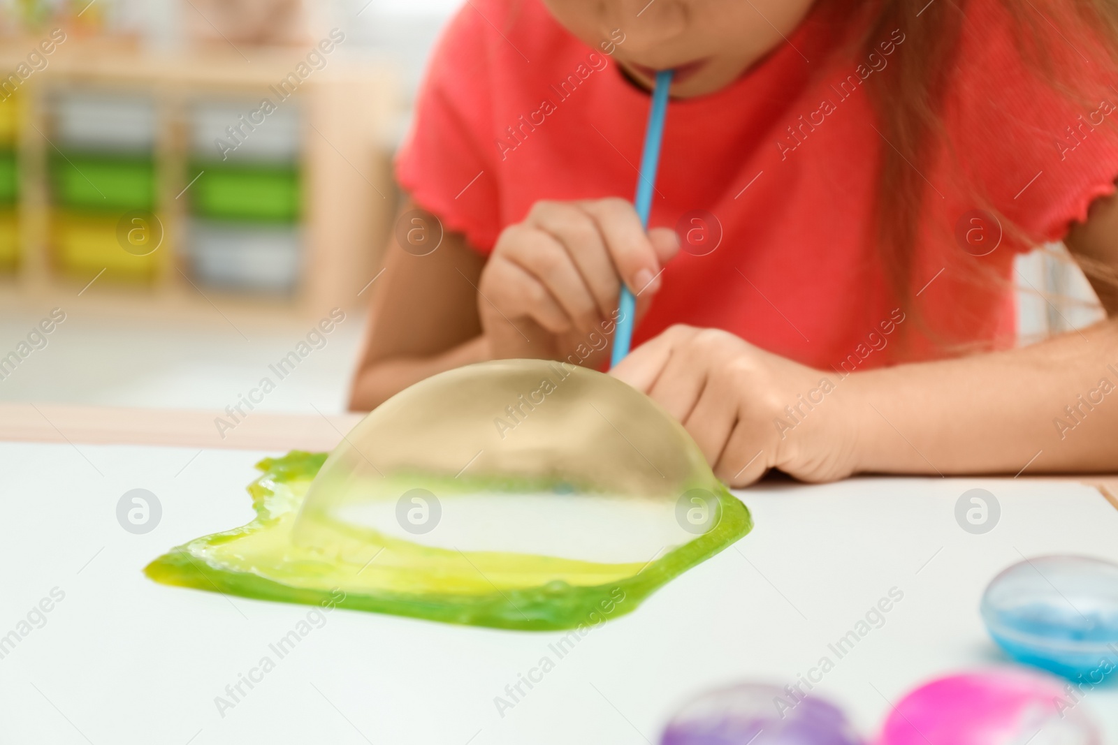 Photo of Little girl blowing slime bubble on white table indoors, closeup