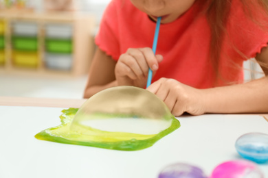 Photo of Little girl blowing slime bubble on white table indoors, closeup