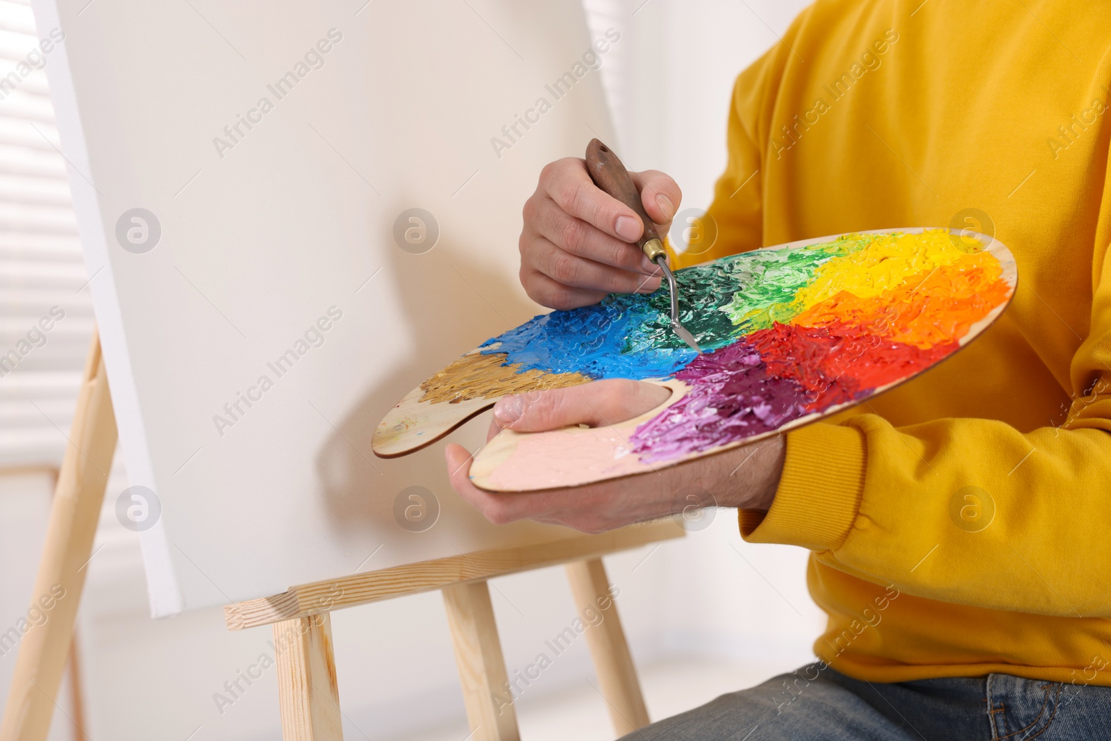 Photo of Man mixing paints on palette with brush in studio, closeup. Using easel to hold canvas