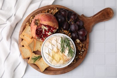 Photo of Board with tasty baked camembert, croutons, grapes, walnuts and pomegranate on white tiled table, top view