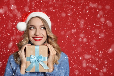 Happy young woman wearing Santa hat with Christmas gift on red background, space for text