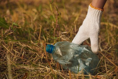 Photo of Woman in gloves picking crumpled bottle from grass outdoors, closeup. Space for text