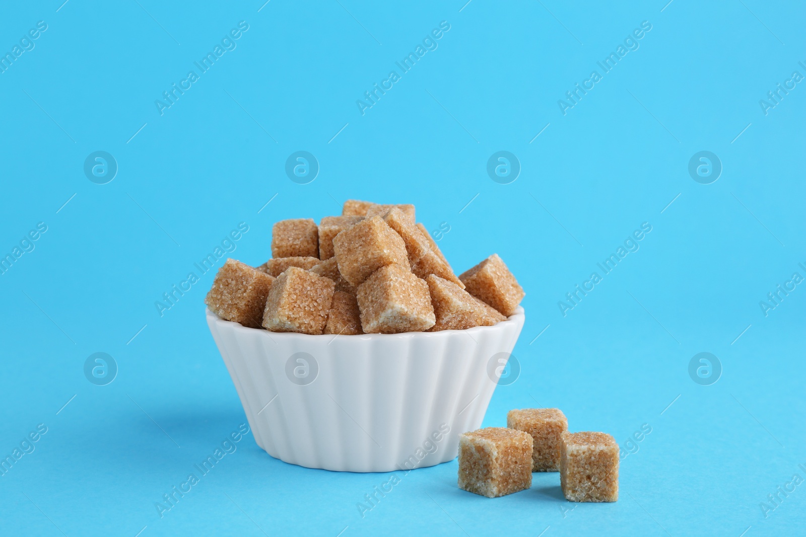 Photo of Brown sugar cubes in bowl on light blue background, closeup