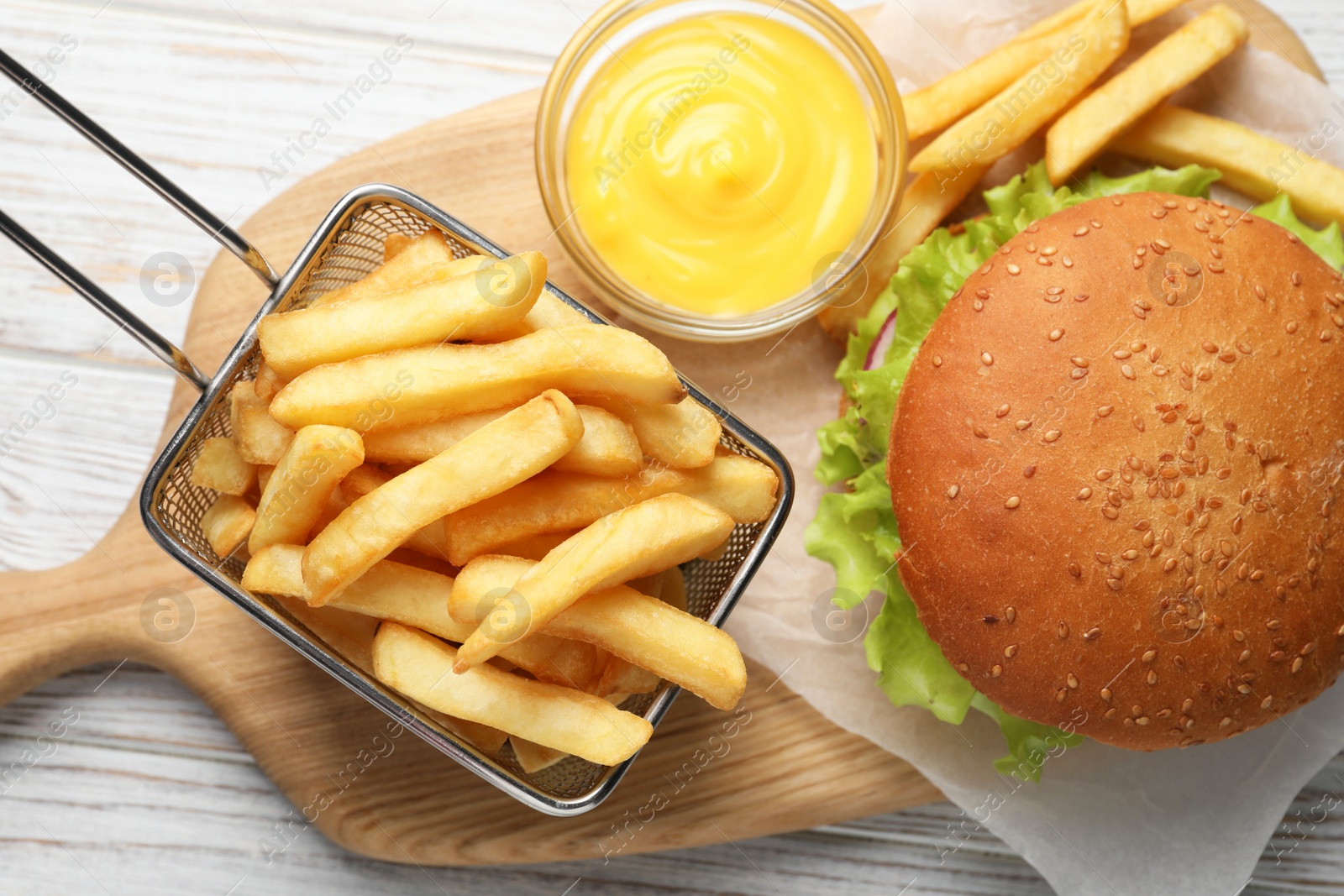 Photo of French fries in frying basket, tasty burger and sauce on white wooden table, flat lay