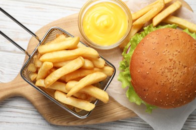 Photo of French fries in frying basket, tasty burger and sauce on white wooden table, flat lay