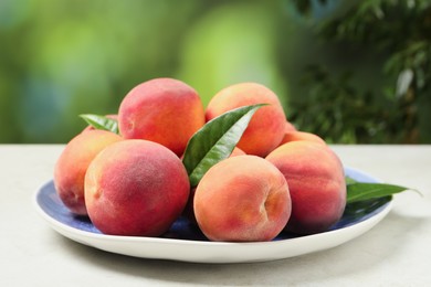 Photo of Many whole fresh ripe peaches and green leaves in plate on white table against blurred background, closeup