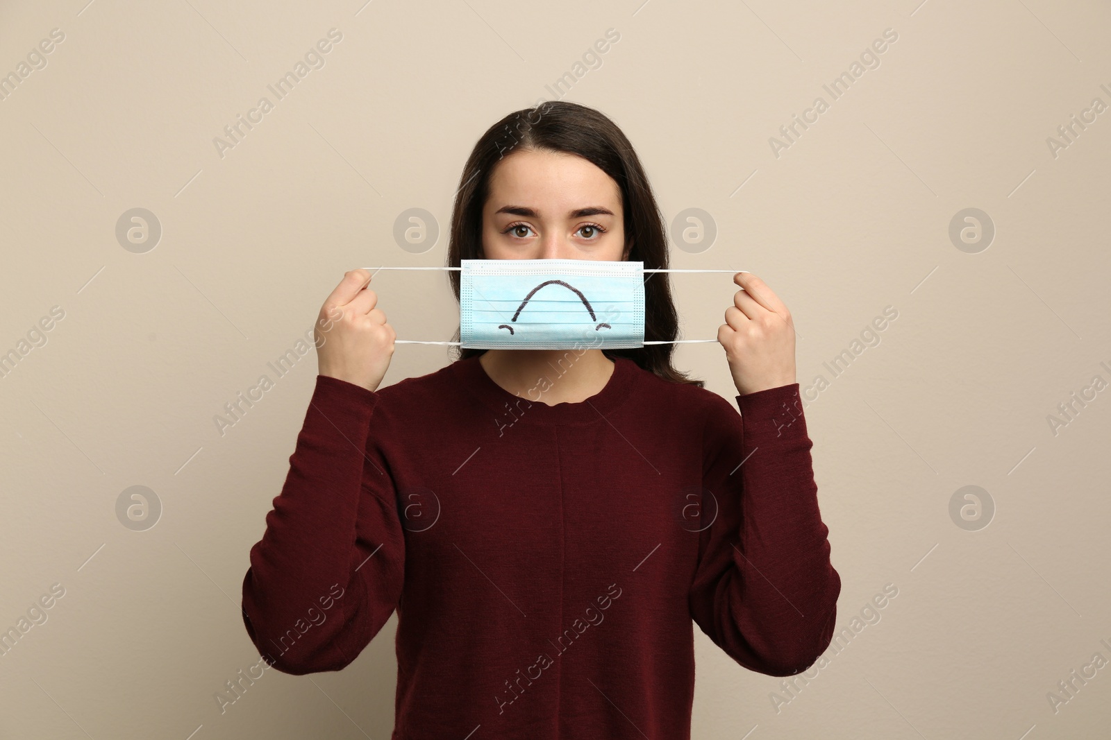 Photo of Woman holding medical mask with drawn sad face on beige background