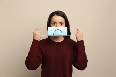 Photo of Woman holding medical mask with drawn sad face on beige background