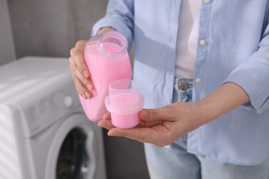 Photo of Woman pouring fabric softener from bottle into cap for washing clothes indoors, closeup