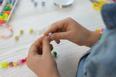 Photo of Little girl making beaded jewelry at white wooden table, closeup