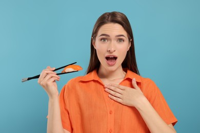 Photo of Emotional young woman holding sushi with chopsticks on light blue background