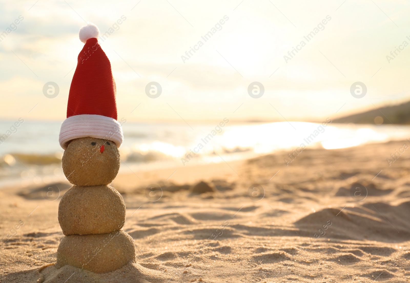 Photo of Snowman made of sand with Santa hat on beach near sea at sunset, space for text. Christmas vacation