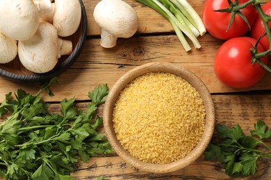 Raw bulgur in bowl, vegetables and parsley on wooden table, flat lay