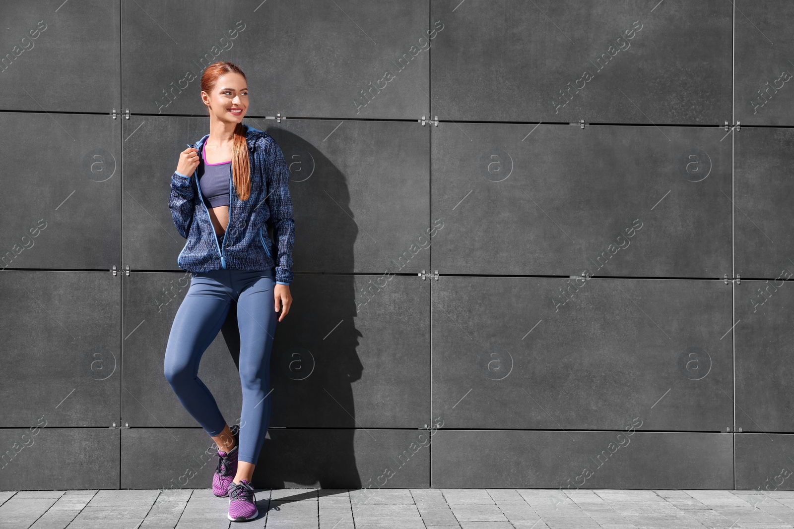 Photo of Beautiful woman in stylish gym clothes near dark grey wall on street, space for text