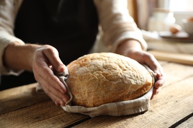 Photo of Man holding loaf of fresh bread at wooden table indoors, closeup