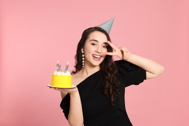 Coming of age party - 21st birthday. Smiling woman showing peace sign and holding delicious cake with number shaped candles on pink background