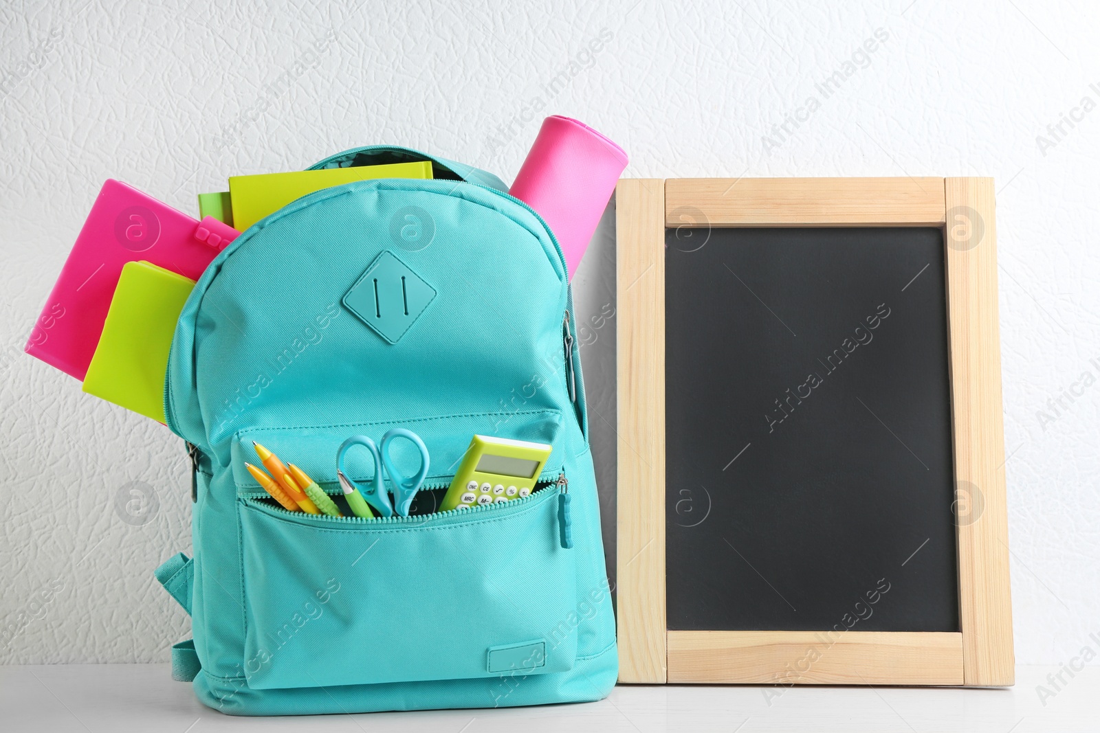 Photo of Blank small chalkboard and backpack with different school stationery on wooden table near white wall. Space for text