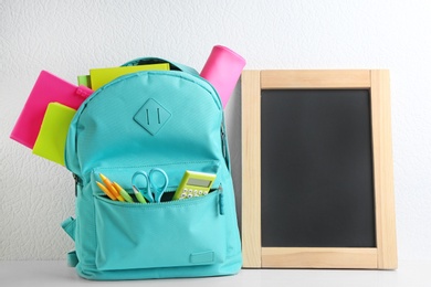 Blank small chalkboard and backpack with different school stationery on wooden table near white wall. Space for text