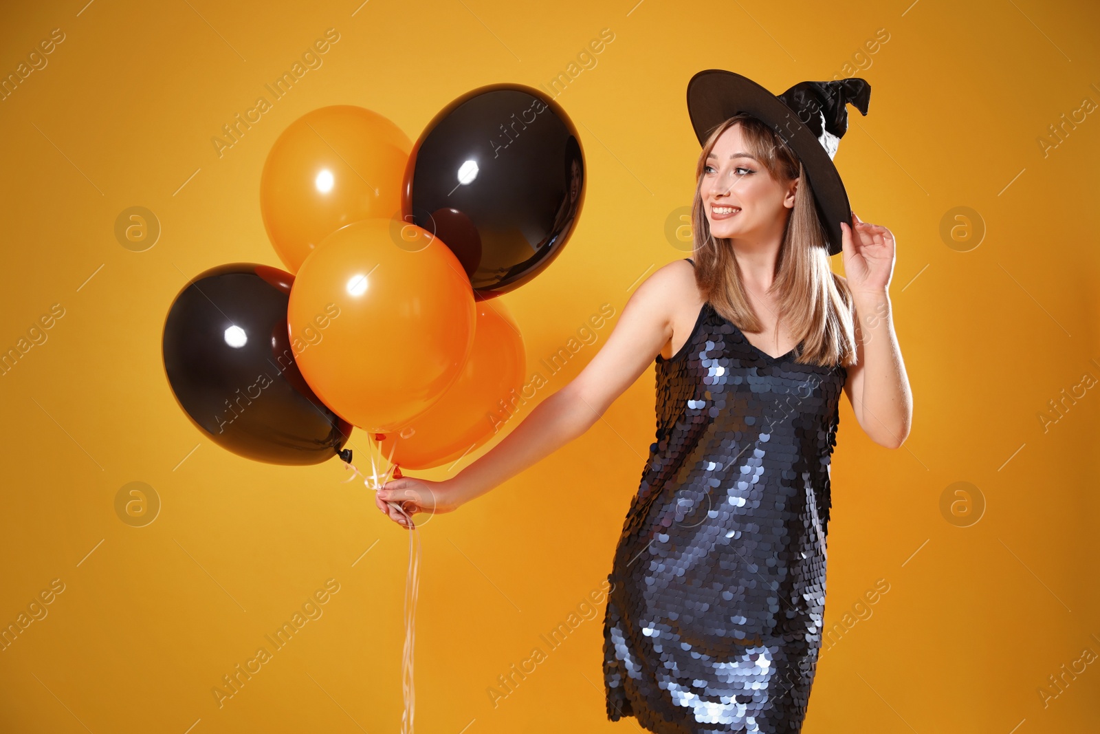 Photo of Beautiful woman in witch costume with balloons on yellow background. Halloween party