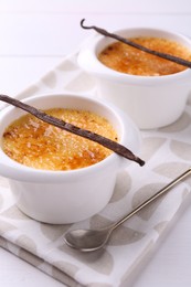 Delicious creme brulee in bowls, vanilla pods and spoon on white table, closeup