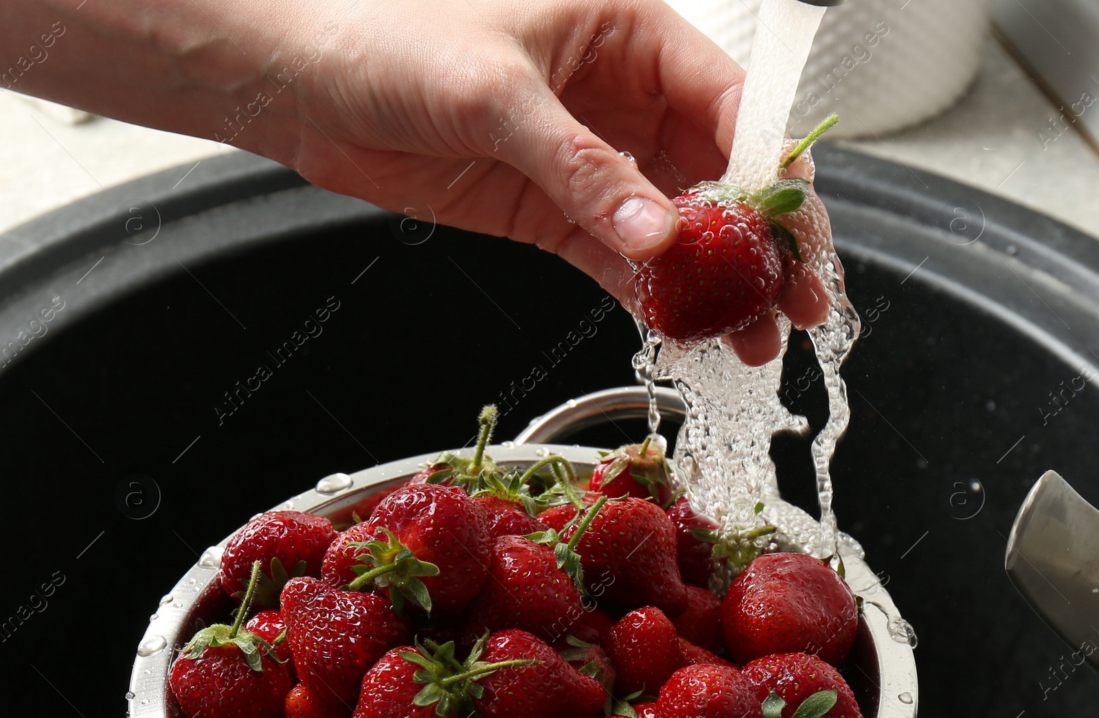 Photo of Woman washing fresh strawberries under tap water in metal colander above sink, closeup