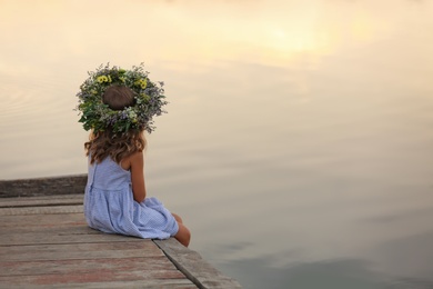 Photo of Cute little girl wearing wreath made of beautiful flowers on pier near river