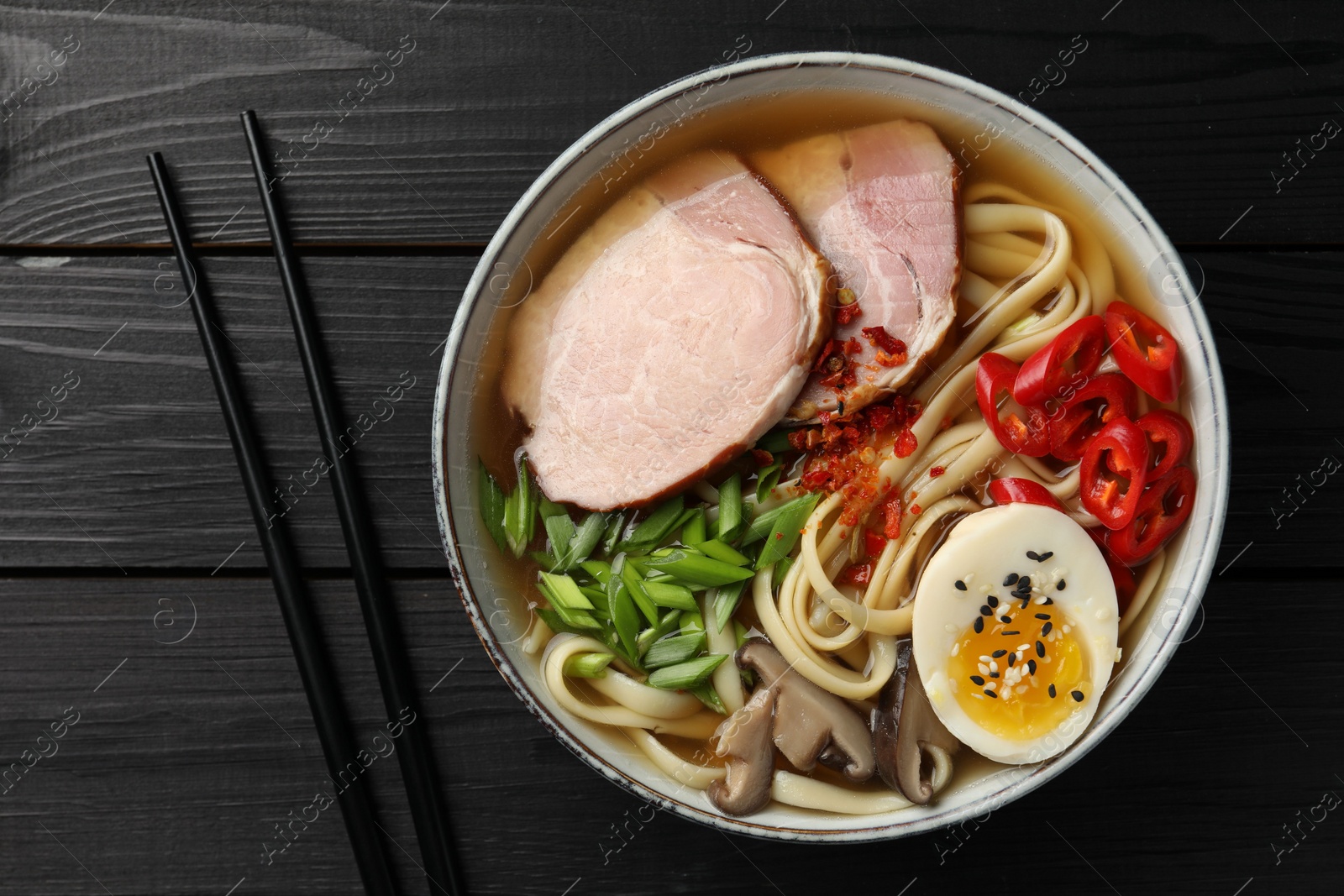 Photo of Delicious ramen in bowl and chopsticks on black wooden table, flat lay. Noodle soup