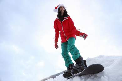 Young snowboarder wearing Santa hat on snowy hill, low angle view. Winter vacation