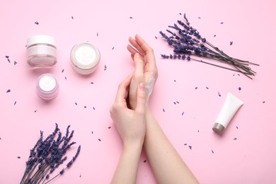 Woman applying hand cream and lavender flowers on pink background, top view