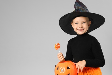 Photo of Cute little girl with pumpkin candy bucket and lollipop wearing Halloween costume on grey background. Space for text