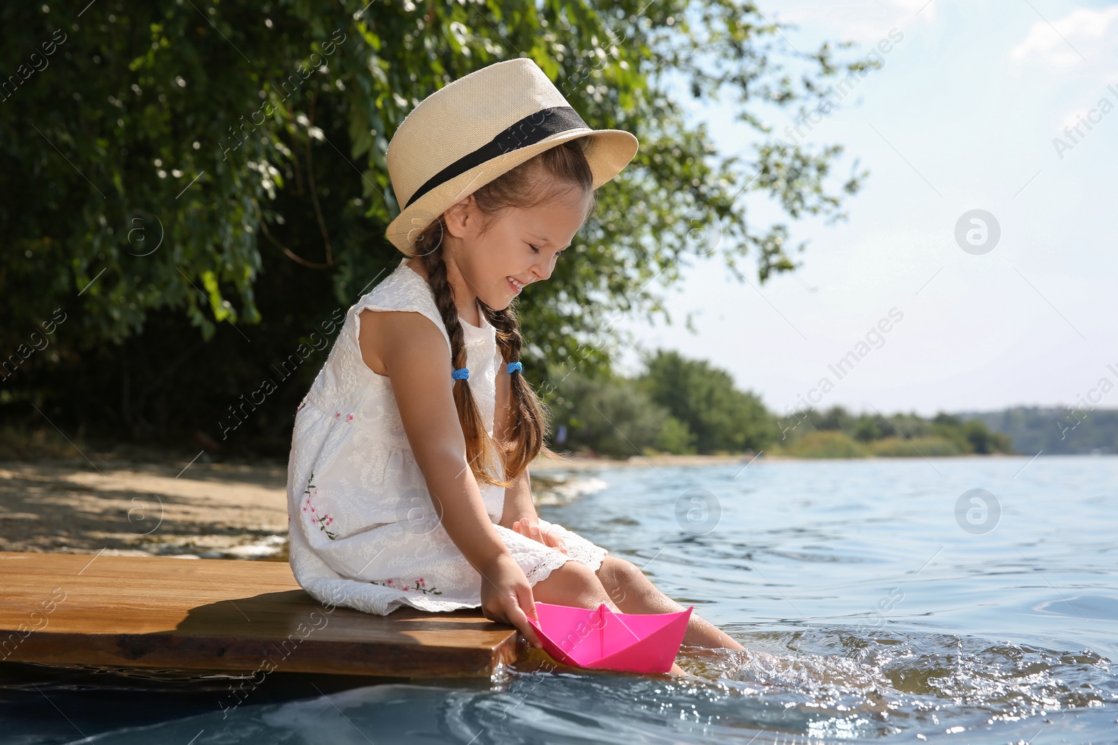 Photo of Cute little girl playing with paper boat on wooden pier near river