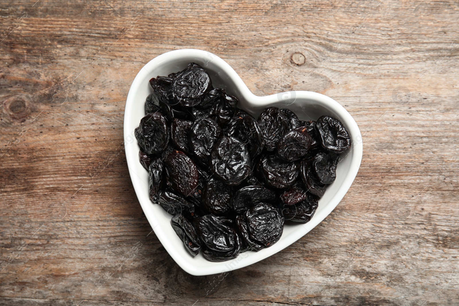 Photo of Heart shaped bowl of sweet dried plums on wooden background, top view. Healthy fruit