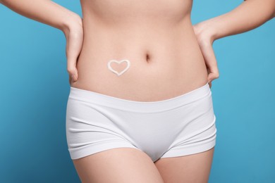 Photo of Woman with heart made of body cream on her belly against light blue background, closeup