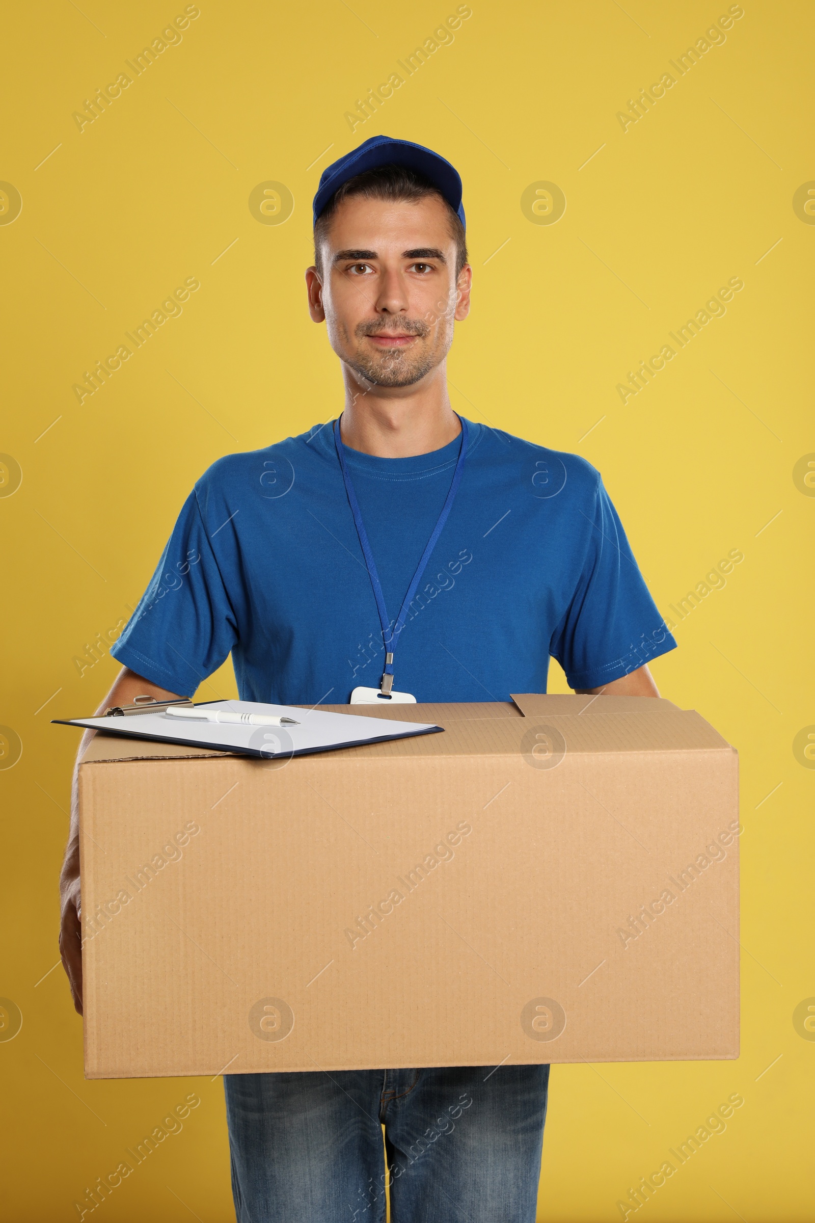 Photo of Happy young courier with cardboard box and clipboard on yellow background