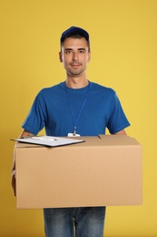 Happy young courier with cardboard box and clipboard on yellow background