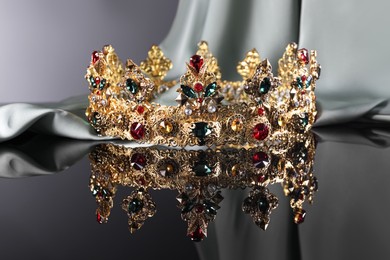 Photo of Beautiful golden crown with gems near light cloth on dark mirror surface