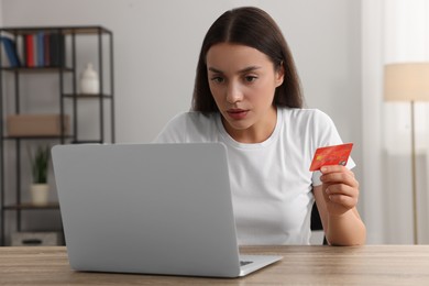 Photo of Confused woman with credit card using laptop at table indoors. Be careful - fraud