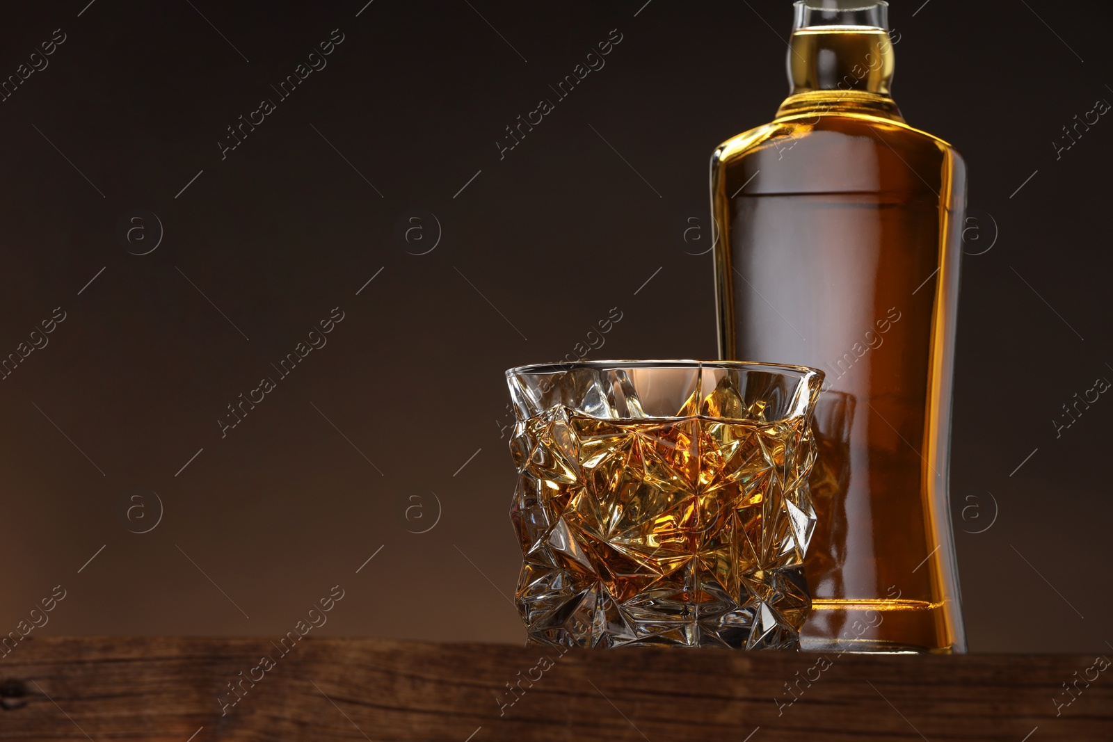 Photo of Whiskey in glass and bottle on wooden table against brown background, space for text