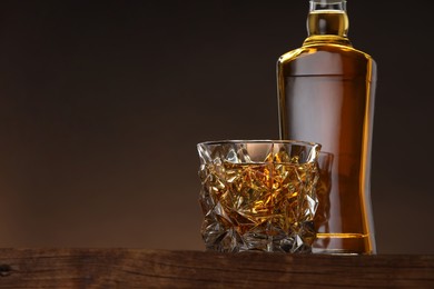 Photo of Whiskey in glass and bottle on wooden table against brown background, space for text