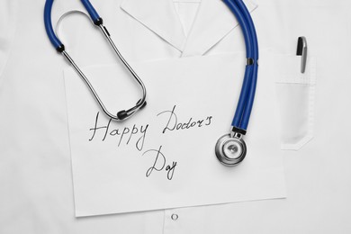 Stethoscope and paper sheet with phrase Happy Doctor's Day on white medical uniform, top view