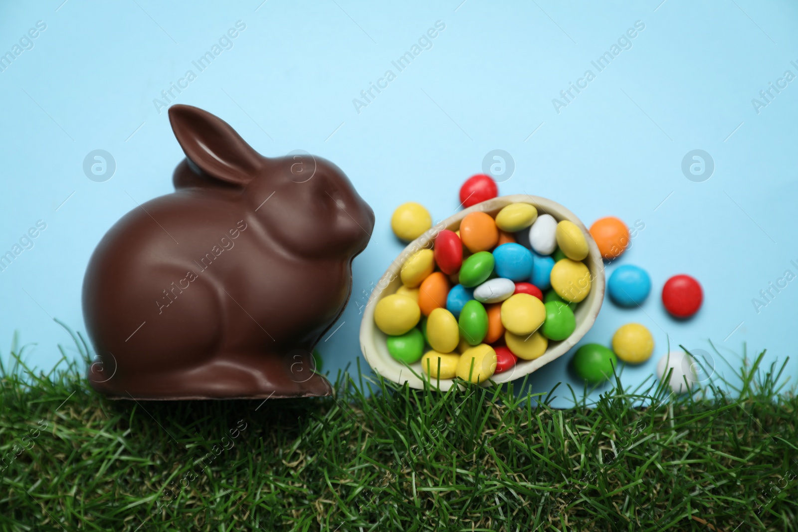 Photo of Chocolate Easter bunny, halved egg and candies with green grass on light blue background