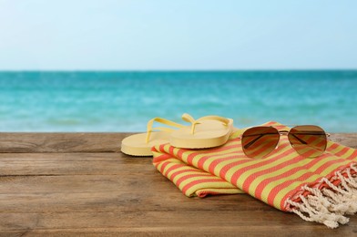Image of Beach towel, flip flops and sunglasses on wooden surface near seashore. Space for text