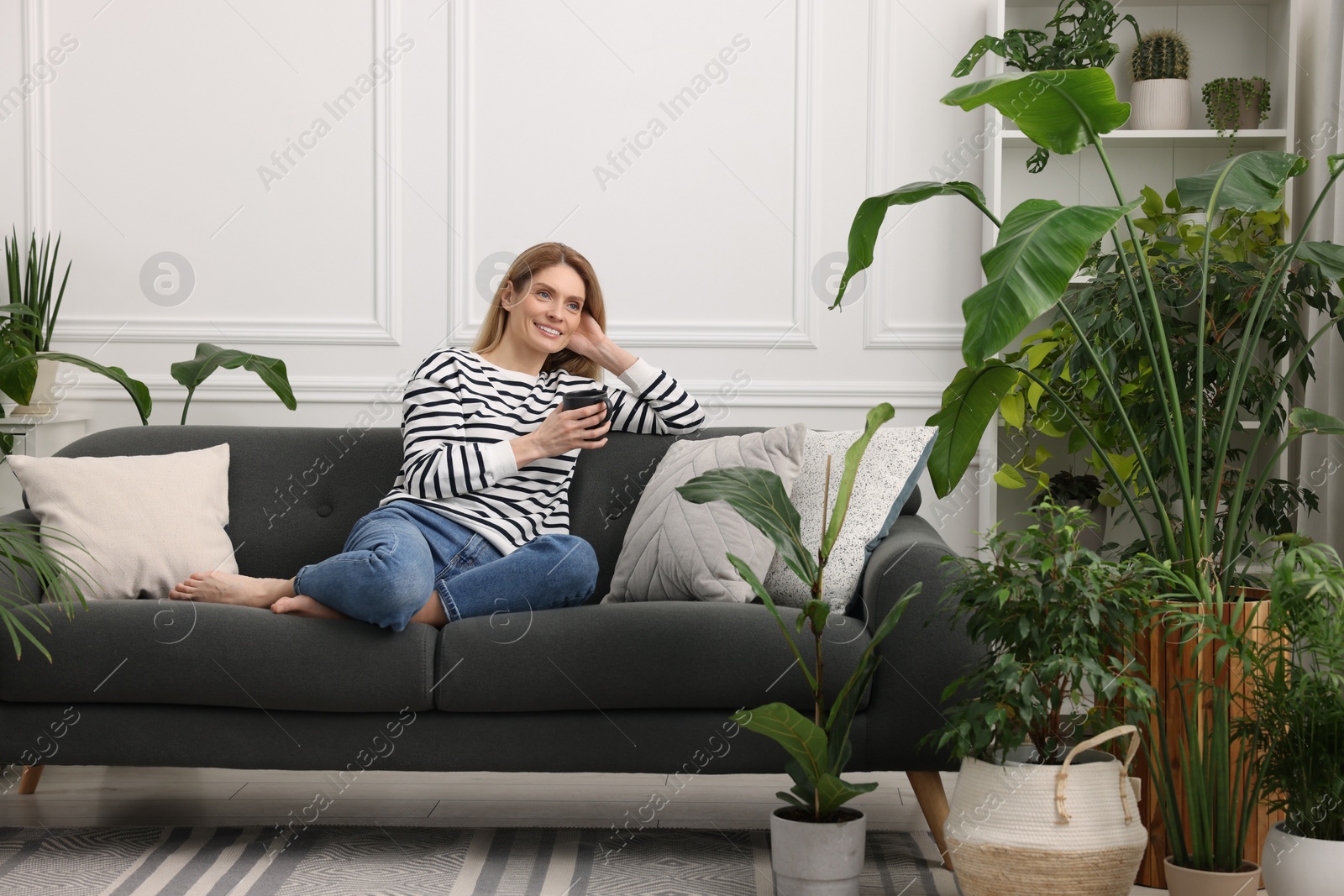 Photo of Woman with cup of drink sitting on sofa surrounded by beautiful potted houseplants at home