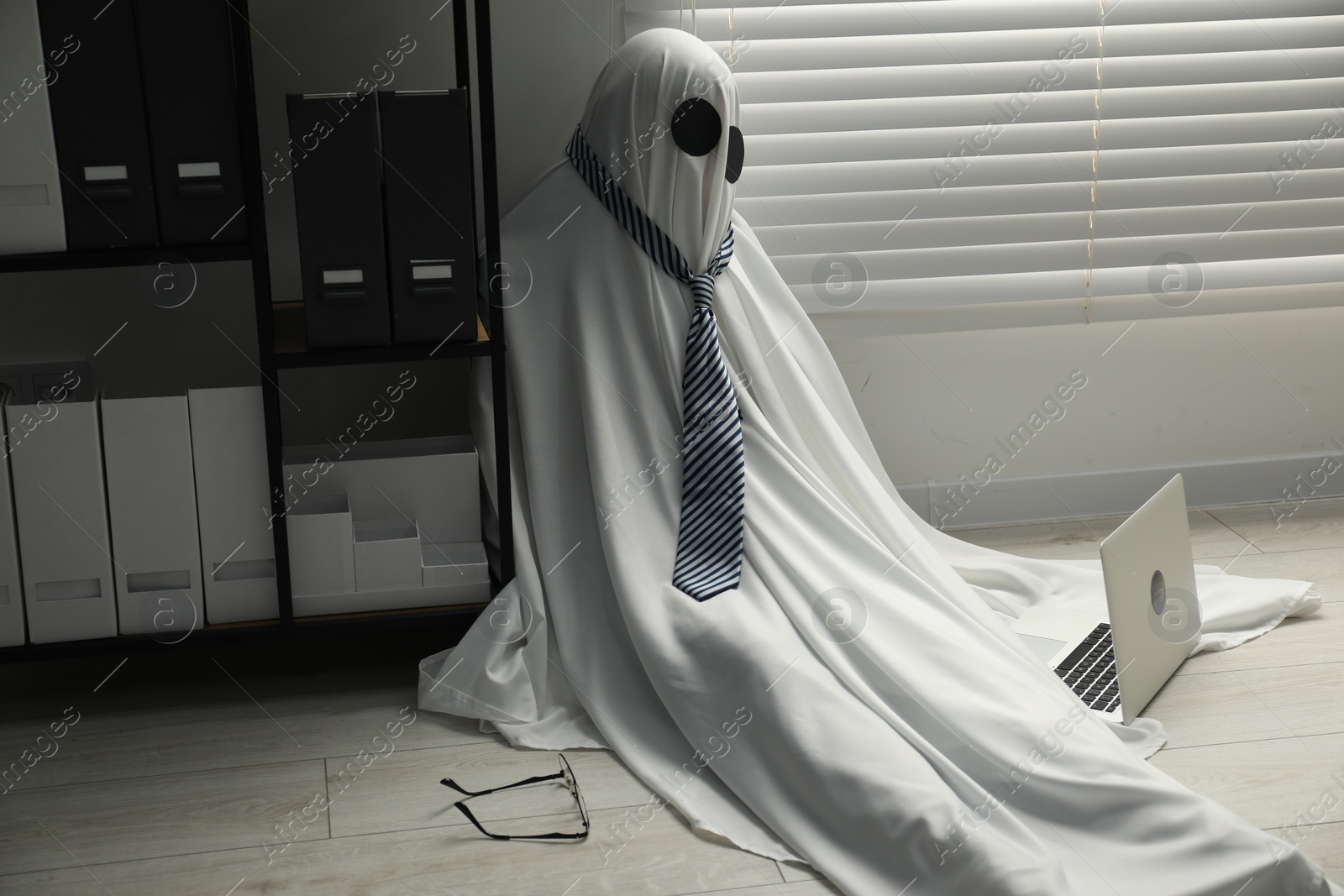 Photo of Overworked ghost. Man in white sheet with laptop on floor in office