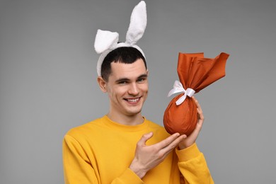 Photo of Easter celebration. Handsome young man with bunny ears holding wrapped gift on grey background