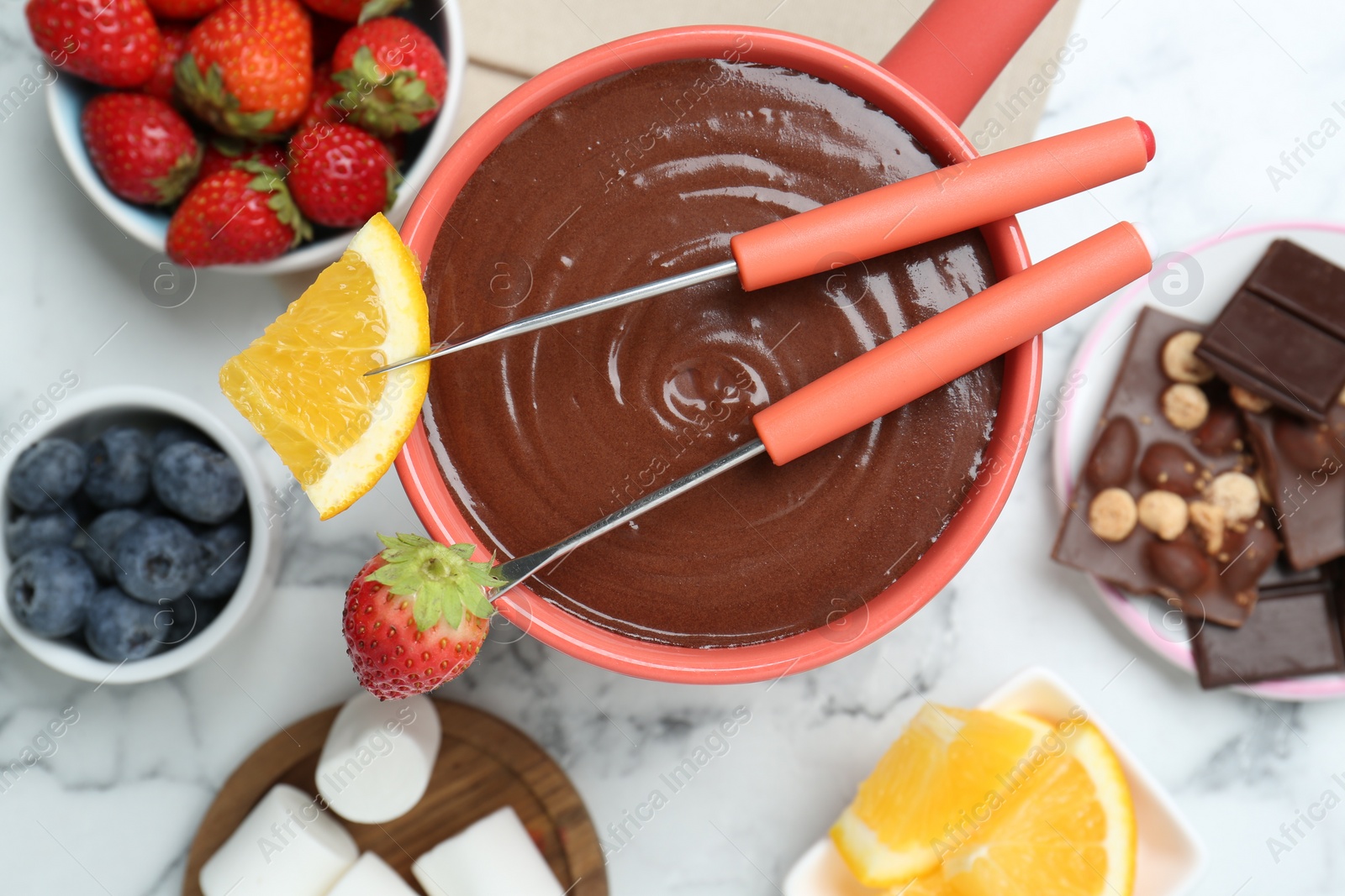 Photo of Fondue pot with melted chocolate, marshmallows, fresh orange, different berries and forks on white marble table, flat lay