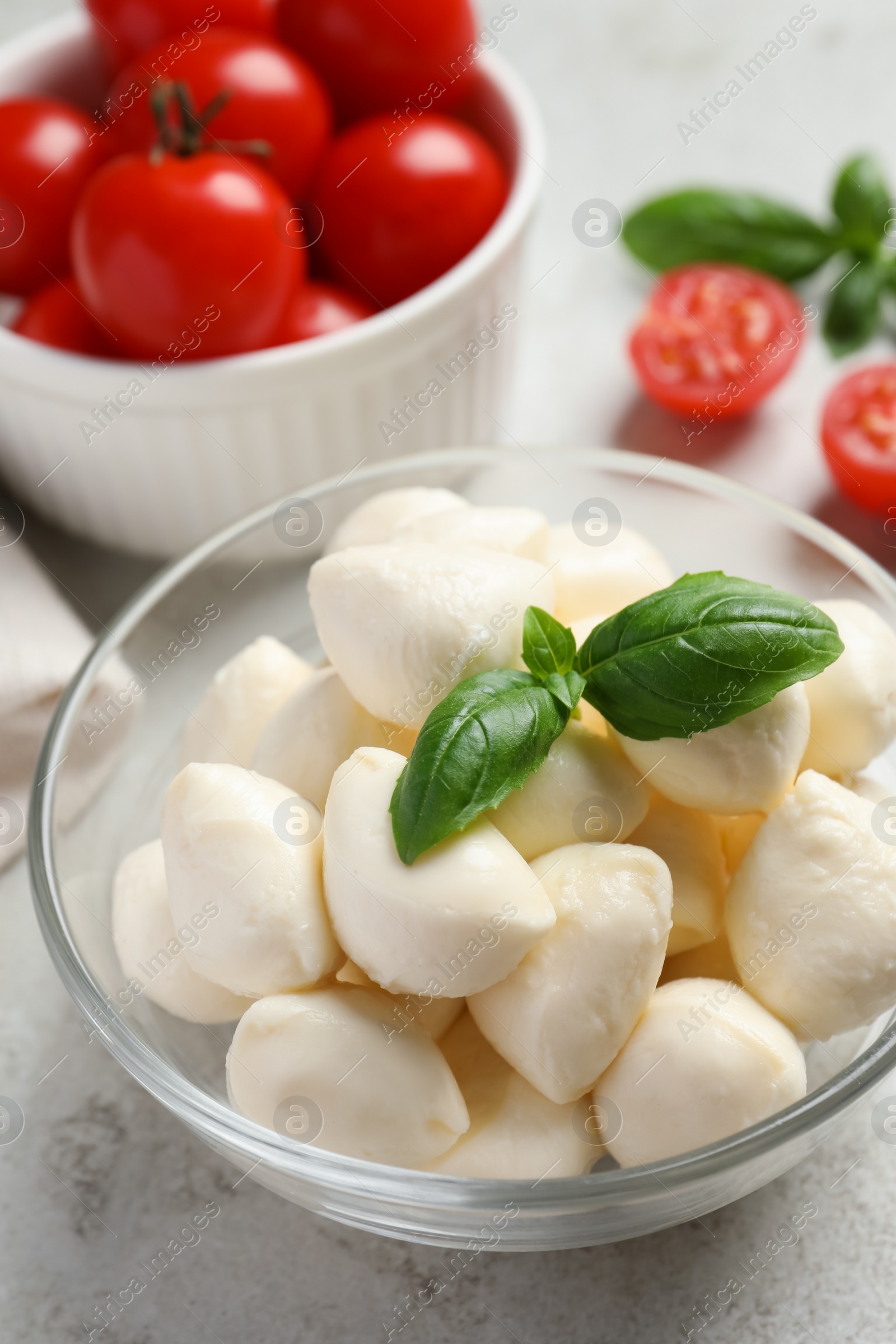 Photo of Delicious mozzarella balls and basil leaves in glass bowl on light gray table, closeup